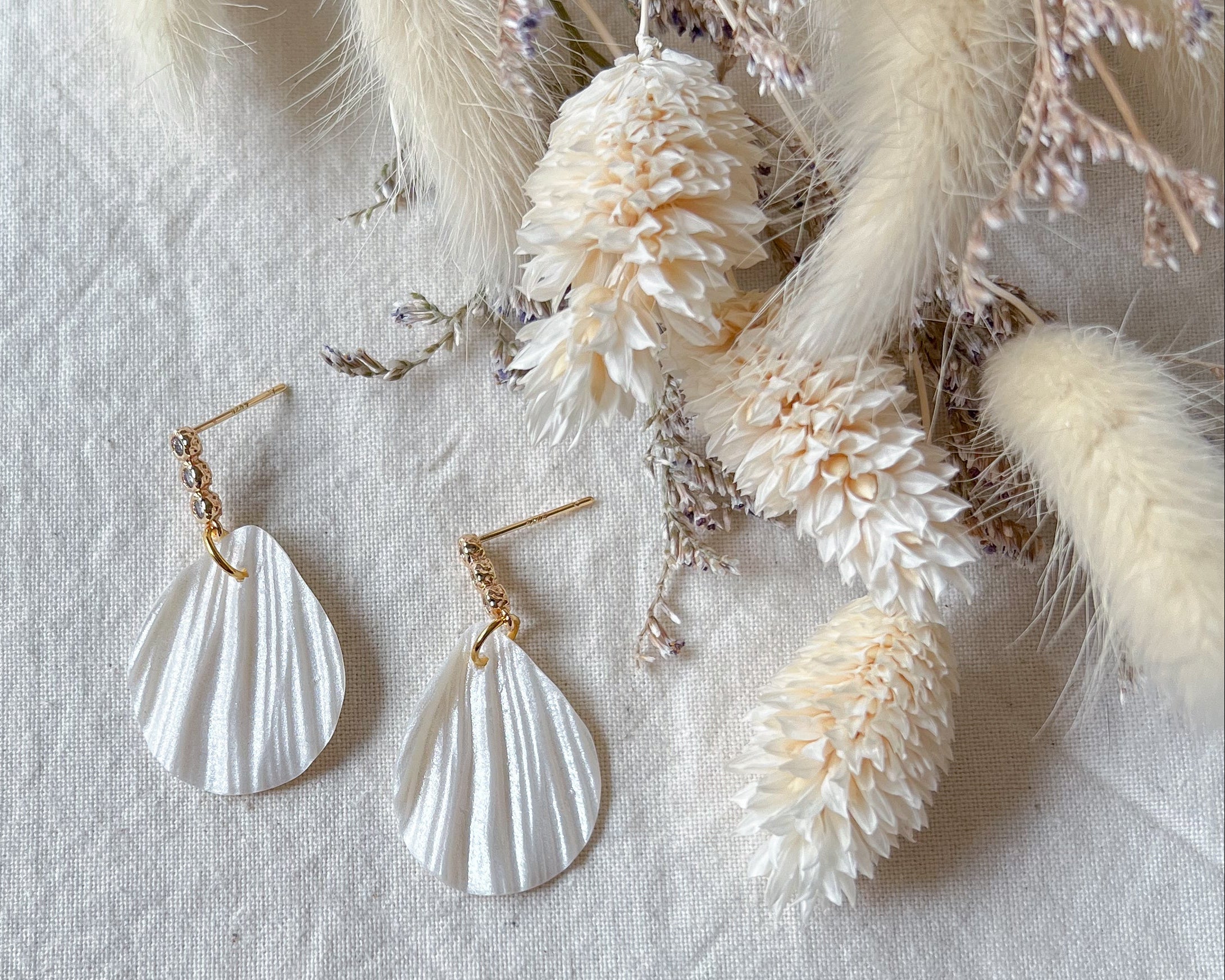 A pair of white, petal-shaped stud earrings with cubic zirconia accents, displayed on a textured fabric next to some dried flowers.
